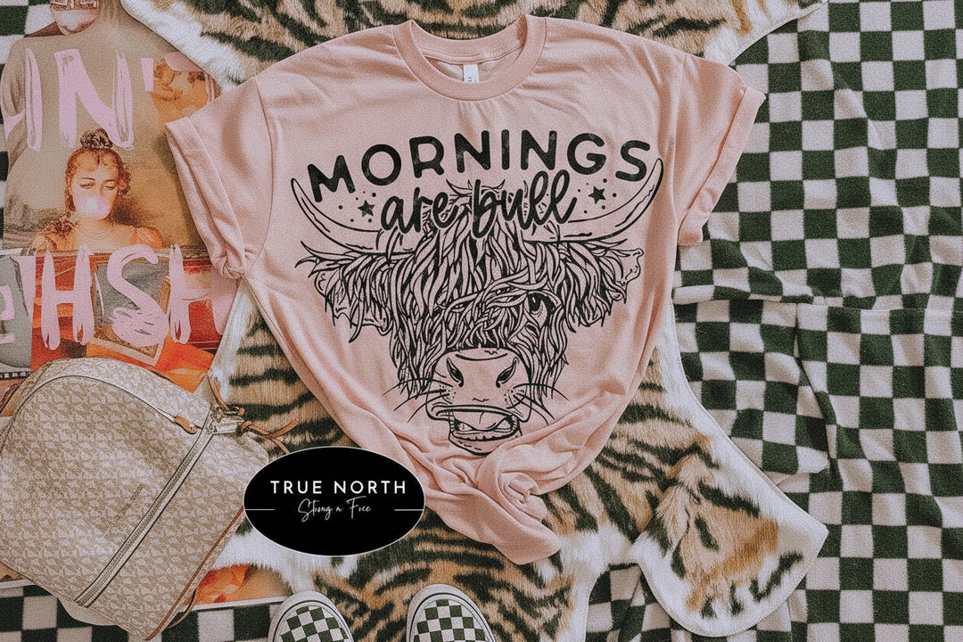 A Blush graphic T-shirt laid flat on a tiger-print faux fur blanket, with a black line-art Highland cow and the words “MORNINGS are bull” printed on the front. A beige patterned handbag, magazine pages, and checkerboard sneakers are styled around it.