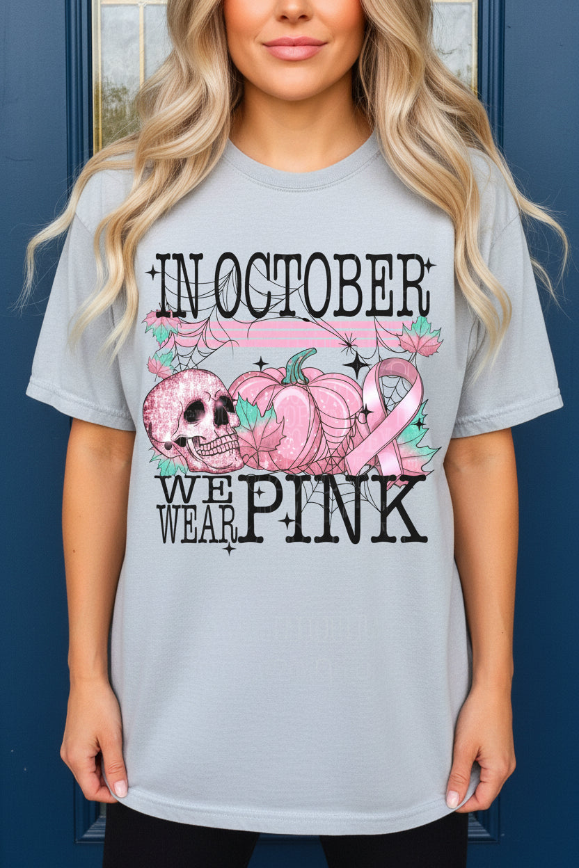 A close-up of a person wearing a grey t-shirt with a large pink and black graphic on the front. The design features the text 'IN OCTOBER WE WEAR PINK' with pumpkins, a skull, spiderwebs, and a pink awareness ribbon.