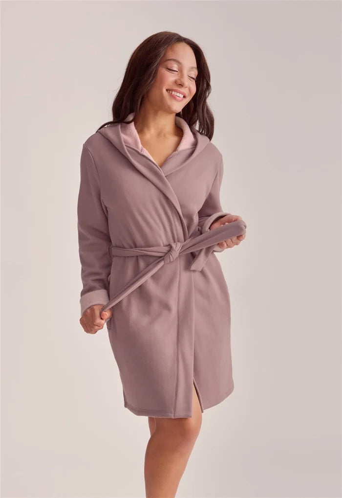 A woman wearing a dusty mauve hooded robe with a matching tie belt, standing against a plain light gray studio background.