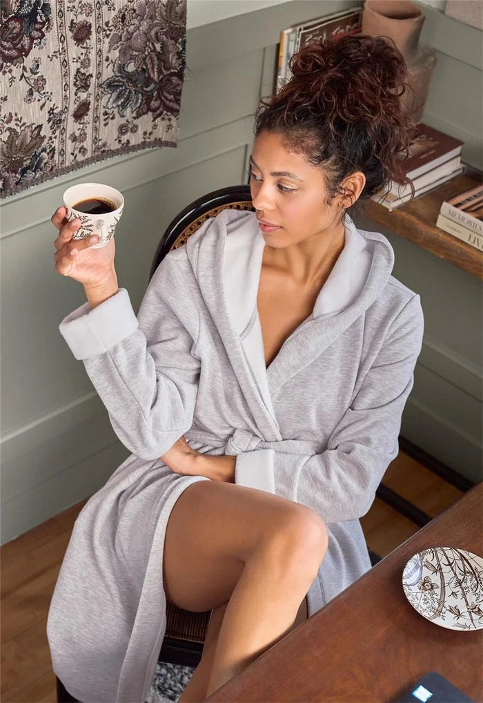 A woman sitting in a chair wearing a light gray plush bathrobe and holding a cup of coffee in a cozy, styled room.