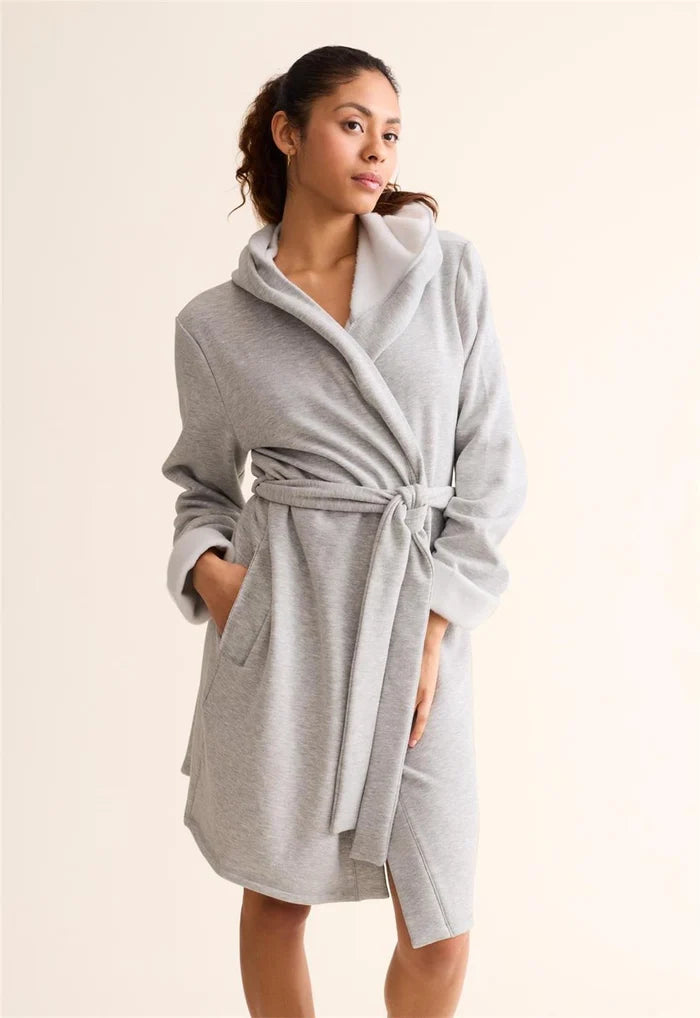 A woman wearing a light gray heathered bathrobe with a shawl collar, tie belt, and front pockets, standing against a plain cream background.