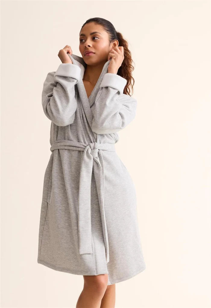 A woman wearing a light gray heathered fleece bathrobe with a tie belt, standing against a solid light beige background.