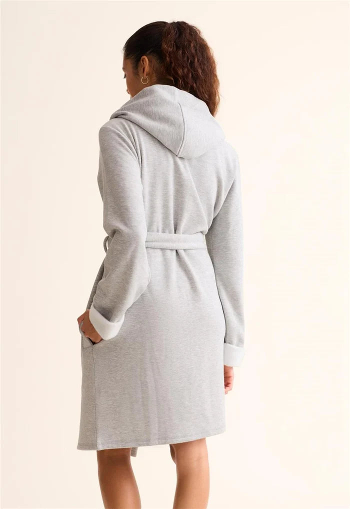 A woman shown from the back wearing a light gray hooded robe with a tie belt, photographed against a plain warm off-white studio background.