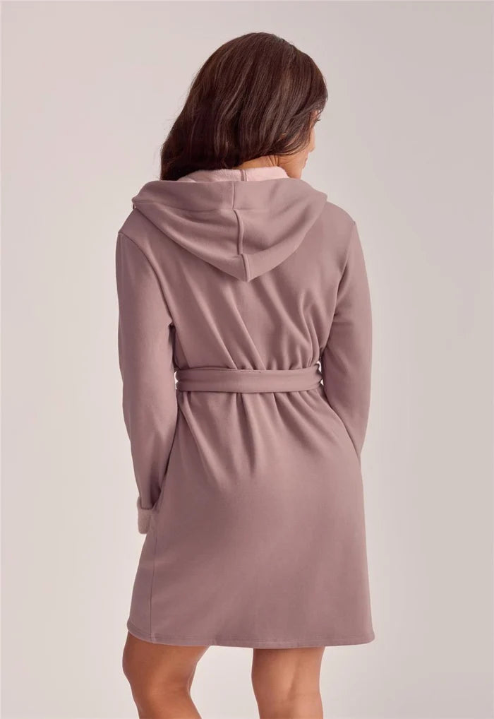 A woman shown from the back wearing a dusty rose hooded robe or dress with a tie belt, photographed against a plain light gray studio background.