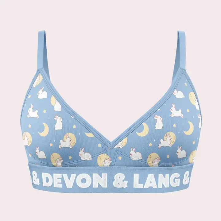 A light blue bra with a repeating pattern of white rabbits, yellow crescent moons, and stars, featuring the brand name '& DEVON & LANG &' printed in white on the underband, set against a pale pink background.