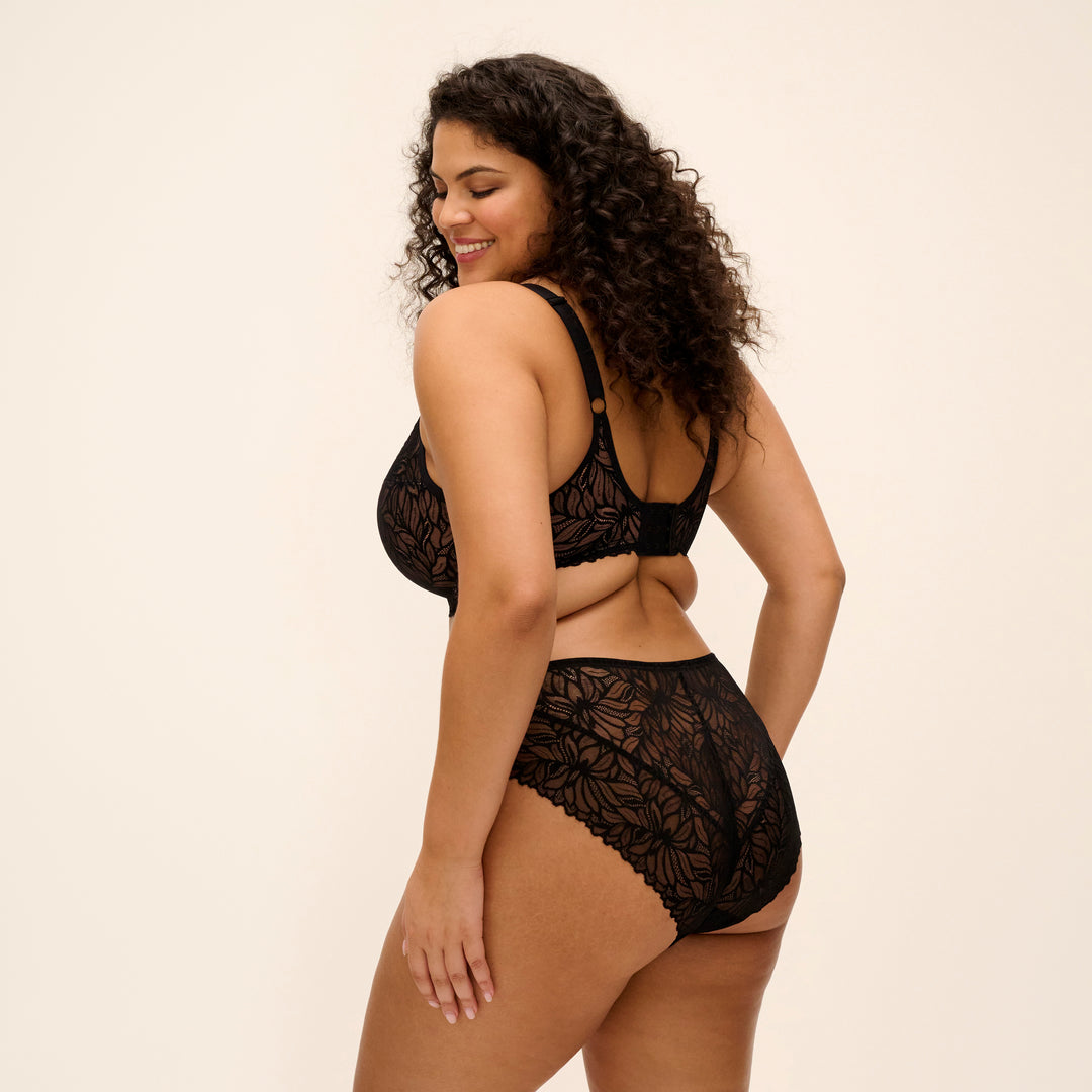A woman modeling a black lace lingerie set, shown from the back-three-quarter view against a plain cream studio background.
