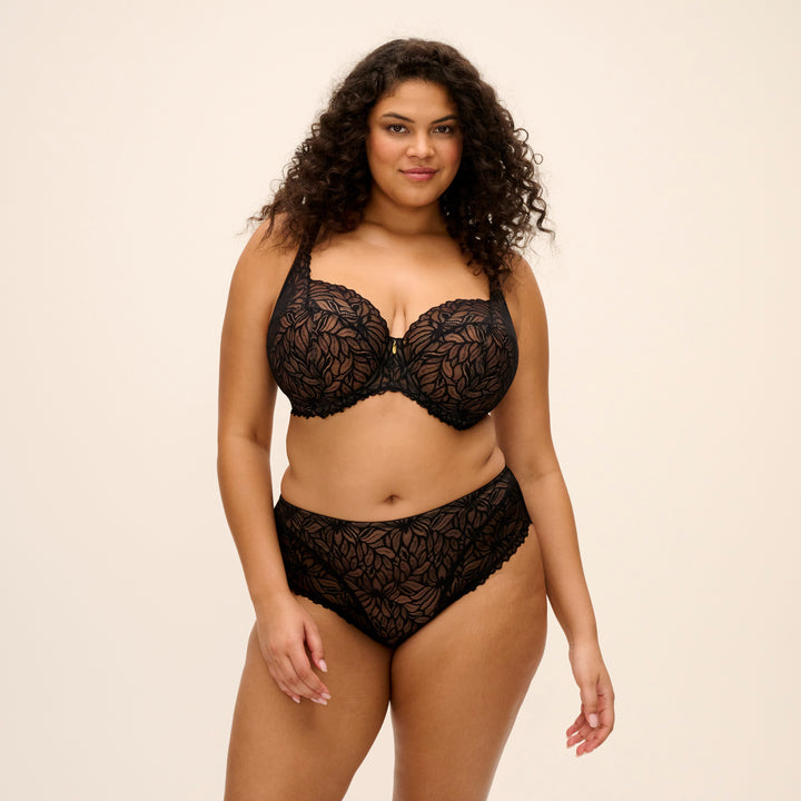 A plus-size model wearing a black lace lingerie set with a floral pattern, featuring a bra and matching high-waisted briefs, against a solid light beige background.