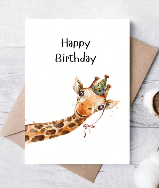 A white 'Happy Birthday' greeting card featuring a watercolor illustration of a giraffe wearing a party hat, styled on top of a brown envelope over a light gray wood-textured surface.