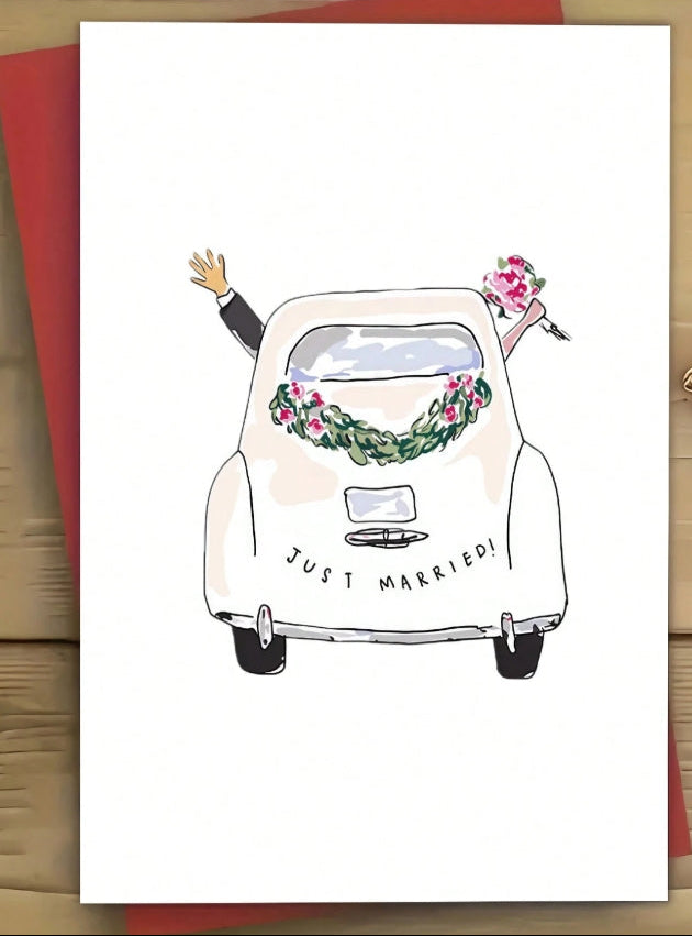 A greeting card featuring a hand-drawn illustration of a cream-colored wedding car with a floral wreath and bouquet, with the words 'JUST MARRIED!' on the front. The card is displayed on a wooden surface with a red envelope behind it.