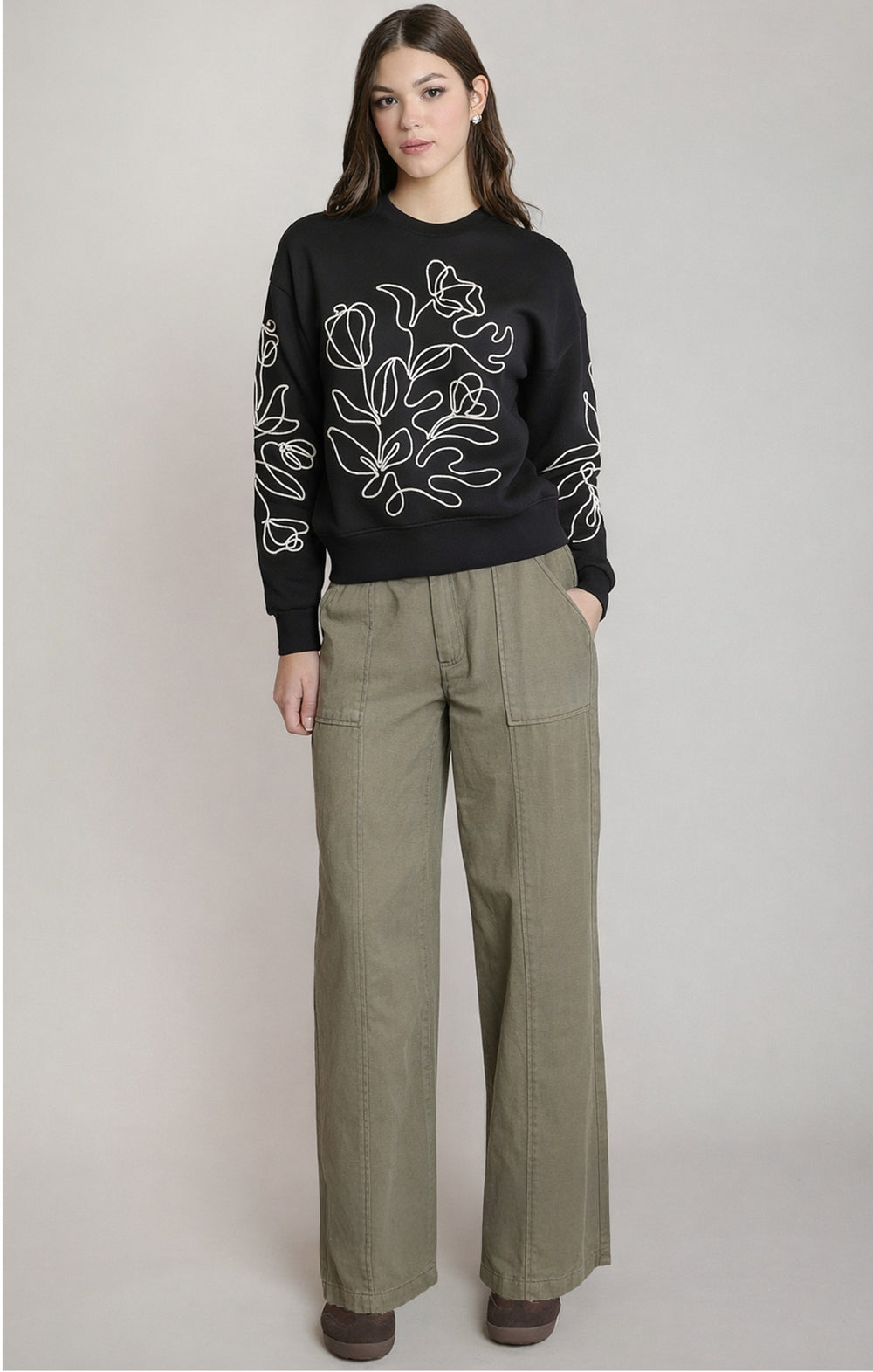 Woman wearing a black sweater with floral patterns and green pants on a light gray background