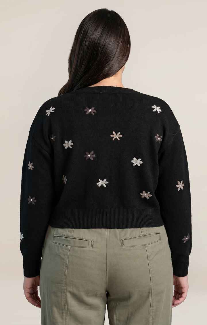 A woman viewed from the back wearing a black knit sweater with small embroidered floral motifs in white and muted pink, paired with olive green pants, against a plain light beige studio background.