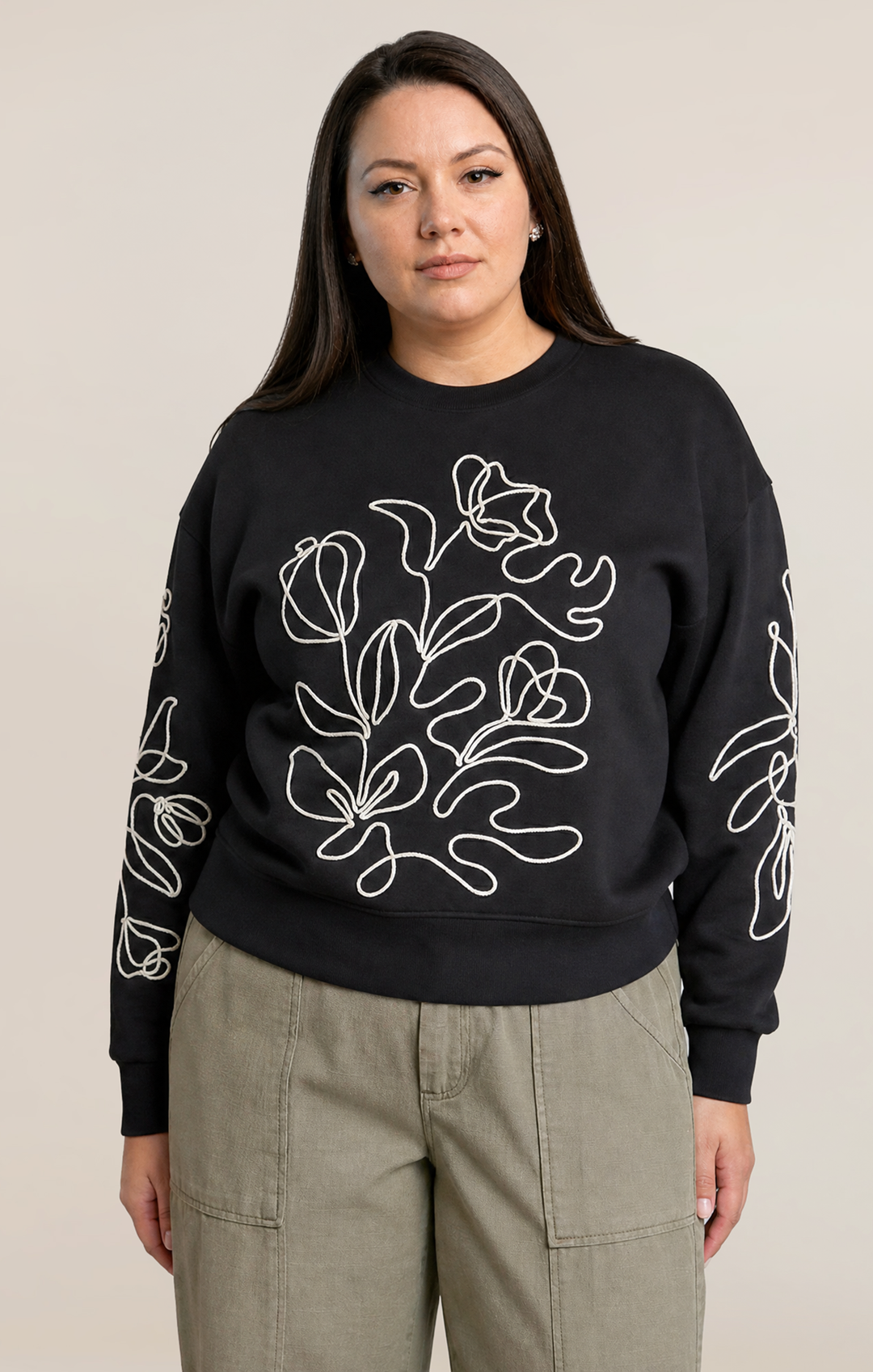 A woman modeling a black crewneck sweatshirt with white line-art floral embroidery on the front and sleeves, paired with olive green cargo pants, against a solid light beige background.