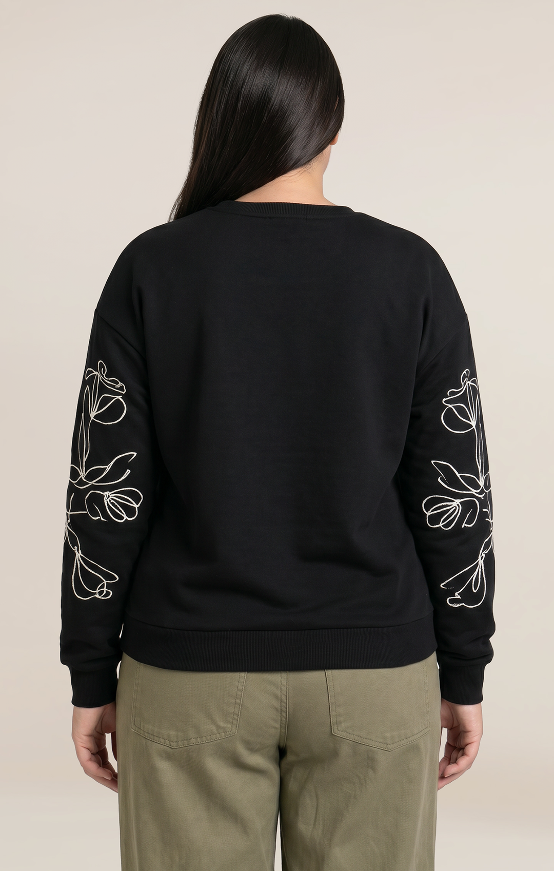 A woman viewed from the back wearing a black crewneck sweatshirt with white floral line-art embroidery on both sleeves, paired with olive green pants, against a solid light beige background.