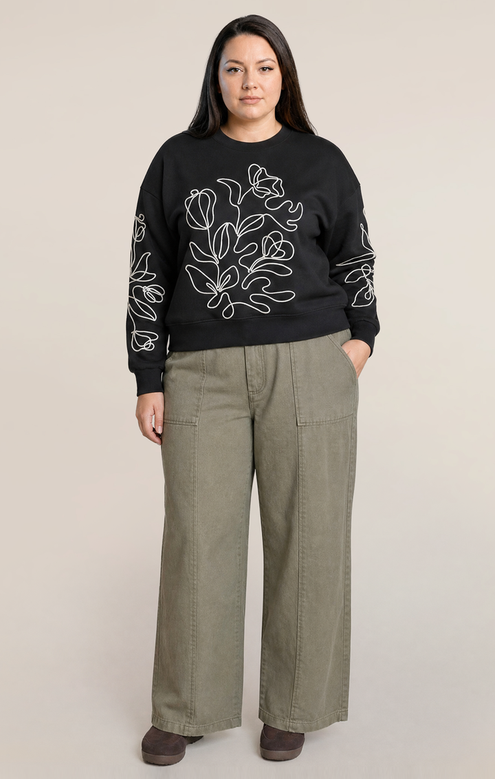 A woman modeling a black crewneck sweatshirt with white floral line-art embroidery on the front and sleeves, paired with olive green wide-leg pants and dark brown shoes, against a solid light beige background.