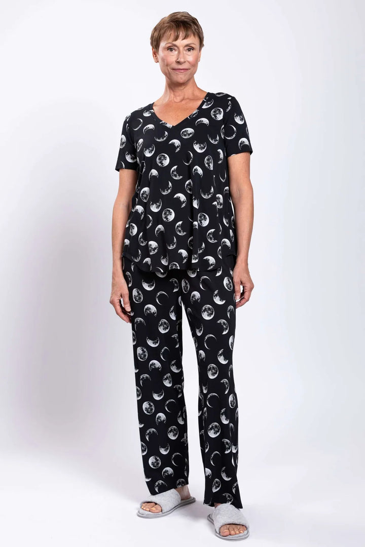 A woman modeling a two-piece pajama set with a black base and a repeating white moon and sun print, paired with light gray slide sandals, against a plain white studio background.