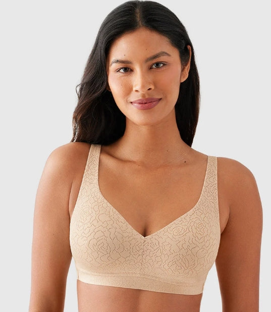 A woman wearing a beige lace underwire bra against a light gray studio background.