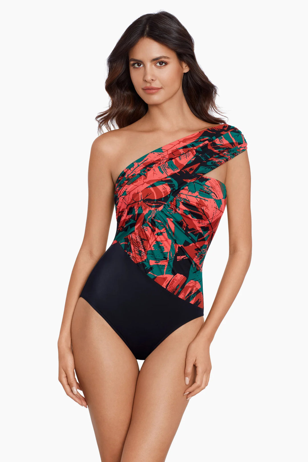 A woman modeling a one-shoulder swimsuit with a red and green tropical leaf print on one side and solid black on the other, against a plain white background.