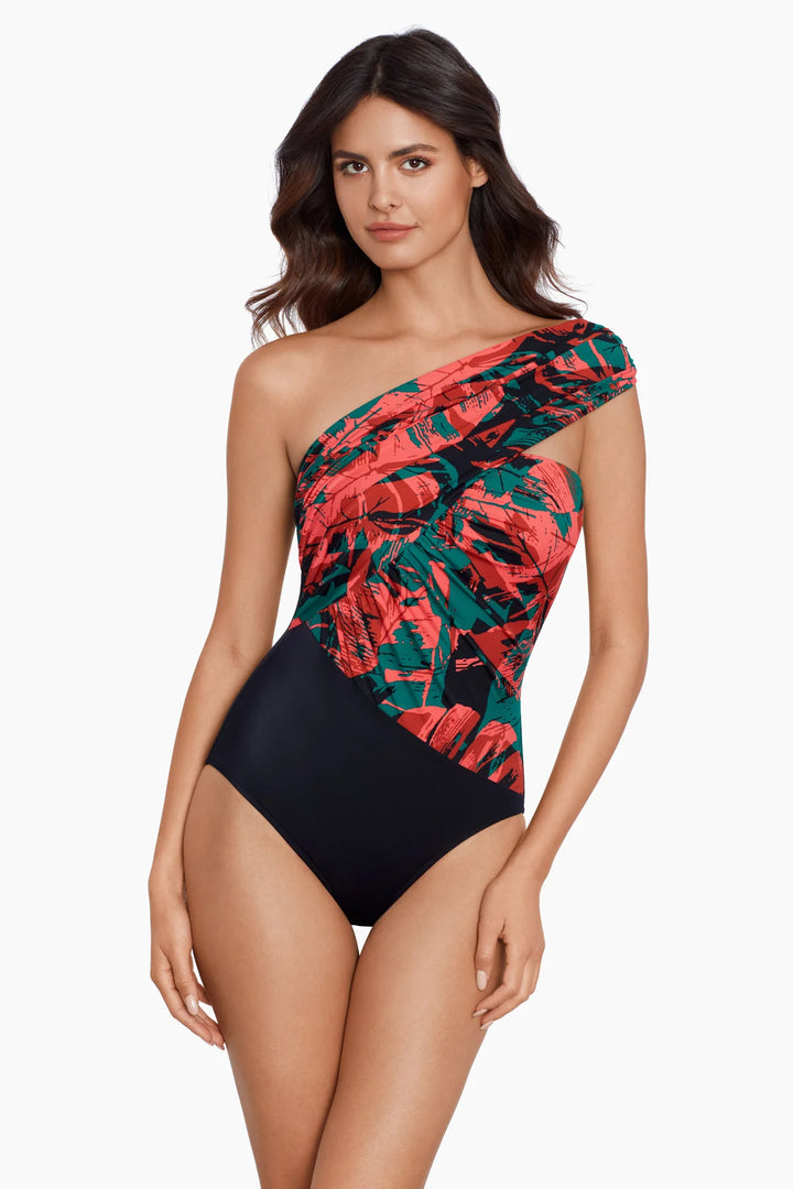 A woman modeling a one-shoulder swimsuit with a red and green tropical leaf print on one side and solid black on the other, against a plain white background.