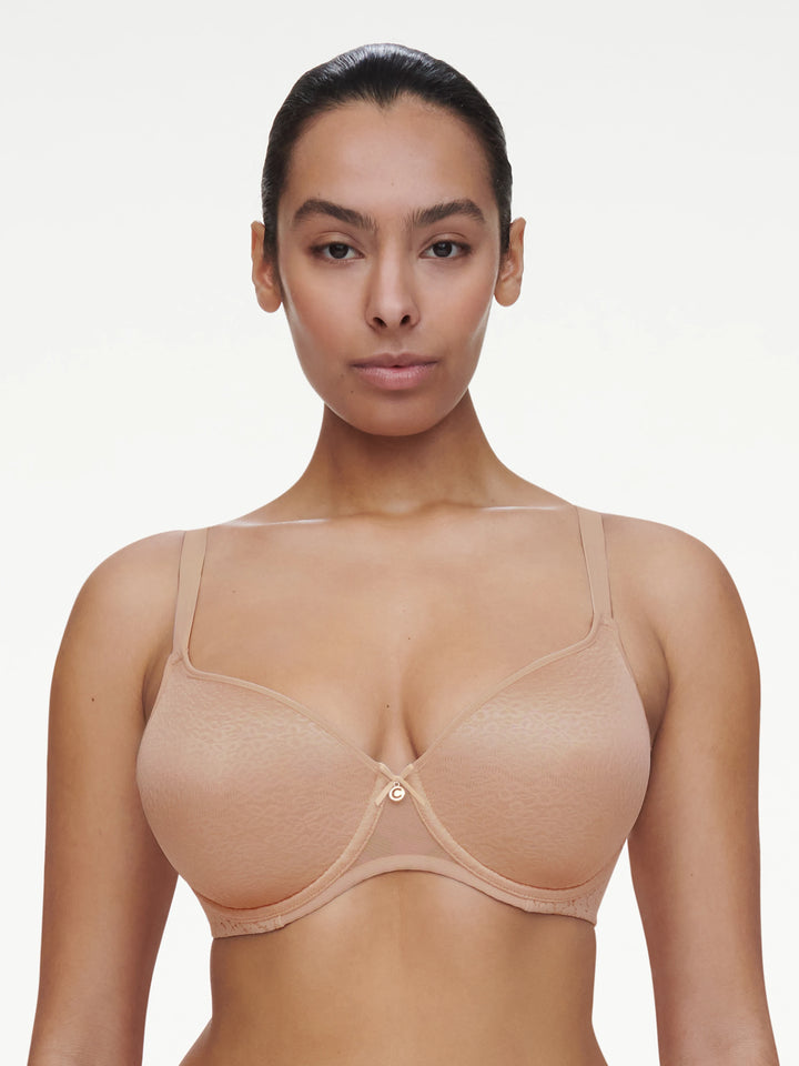 A woman modeling a seamless, bra in a nude beige color, shown from the chest up against a clean, light off-white background.