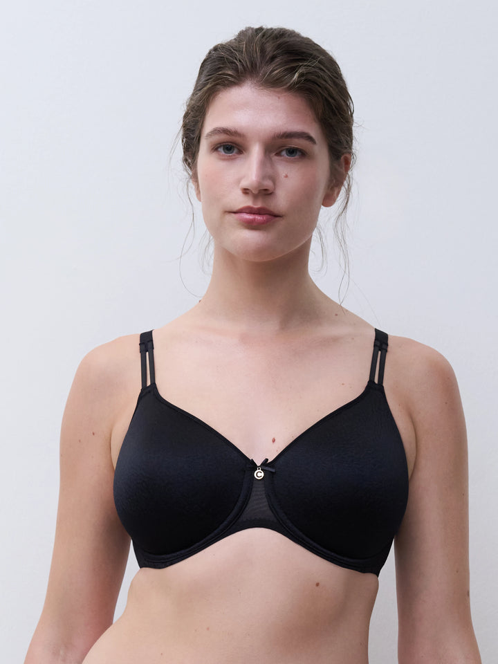 A woman modeling a black seamless underwire bra with double straps and a small gold button closure at the center front, set against a plain light gray background.