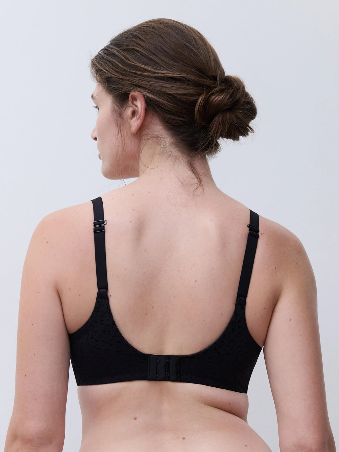 Rear view of a woman wearing a black underwire bra with adjustable shoulder straps and a center back strap, shown against a light gray studio background.