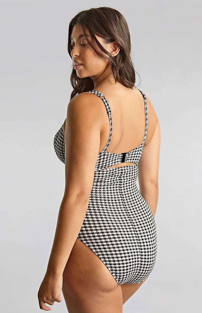A woman modeling a one-piece swimsuit with a black and white houndstooth pattern, shown from a three-quarters back view against a light gray studio background.
