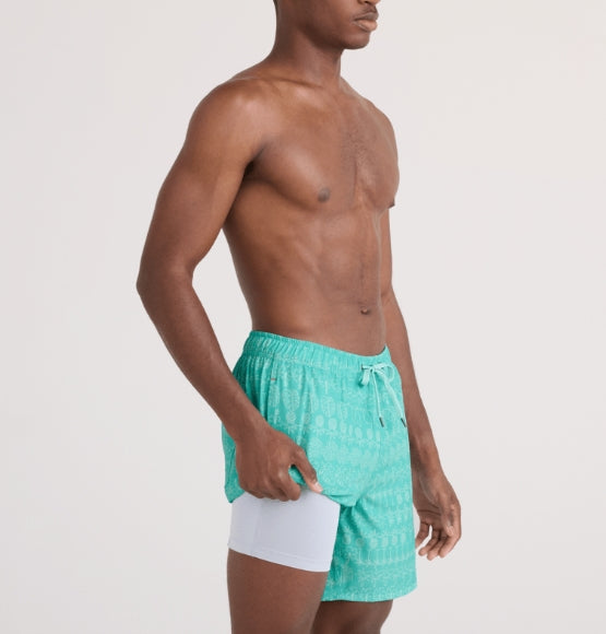 A man wearing turquoise patterned swim trunks with a white under-trunk insert, shown from the waist up against a light gray studio background.