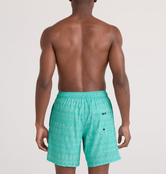A man shown from the back wearing turquoise patterned swim trunks against a light gray studio background.