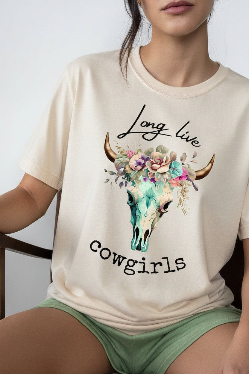 A person wearing a light beigeT-Shirt with a graphic print of a floral cow skull and the text 'Long live cowgirls' in black script. The person is also wearing sage green shorts, seated indoors.