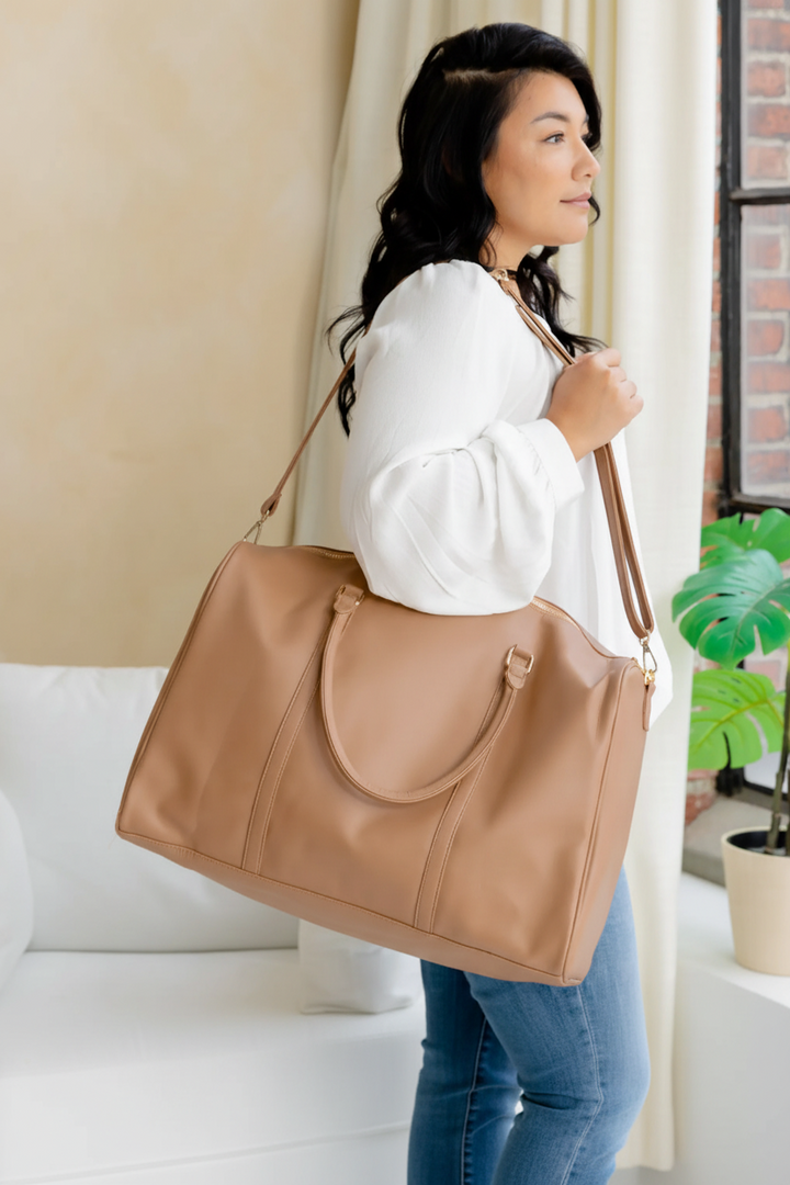 Woman holding a tan handbag with in a room with a plant and window.