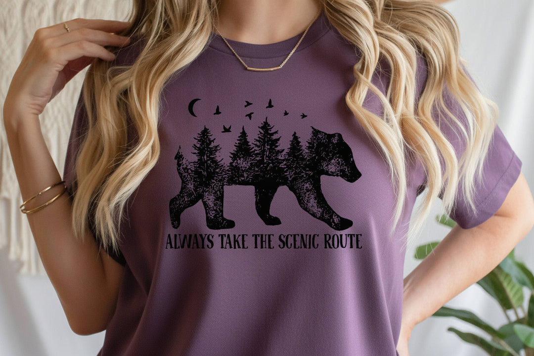 A woman wearing a Purple t-shirt with a black graphic of a bear filled with pine trees and the text 'ALWAYS TAKE THE SCENIC ROUTE'.