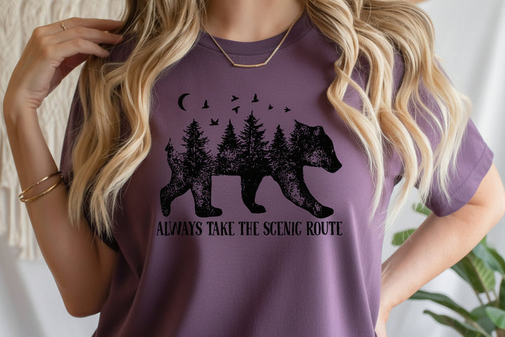 A woman wearing a Purple t-shirt with a black graphic of a bear filled with pine trees and the text 'ALWAYS TAKE THE SCENIC ROUTE'.